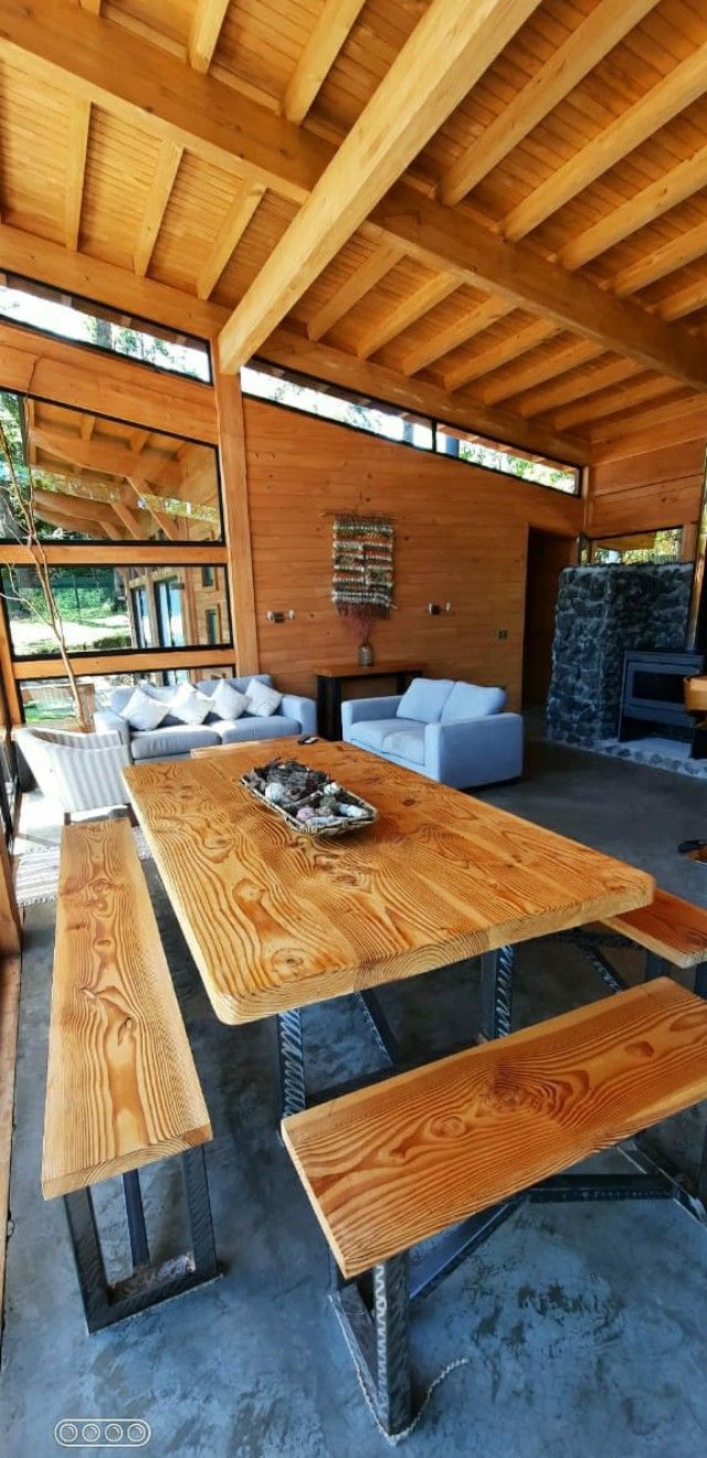 A wooden dining table is surrounded by benches in a spacious open-plan area. Large windows allow natural light to fill the room, revealing views of the surrounding landscape. A cozy seating arrangement with soft cushions is positioned nearby, enhancing the warmth of the space.