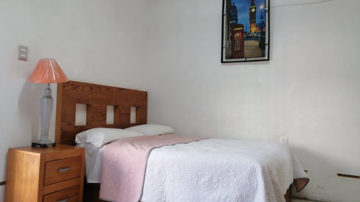 A simple guest room is depicted, featuring a single bed covered with a white blanket. A wooden headboard with cut-out designs complements the space. A bedside table holds a lamp, and a framed picture of a London scene adorns the wall.