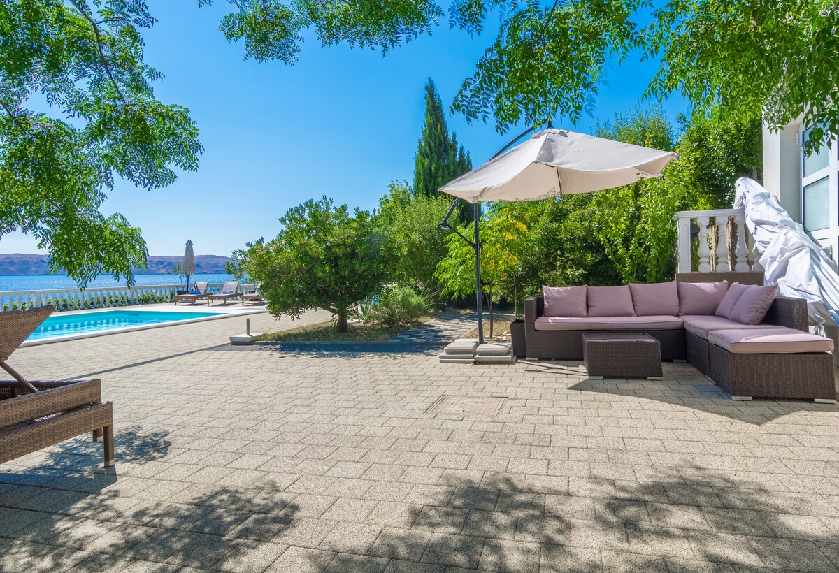 A spacious patio area features a comfortable seating arrangement with soft cushions under a large umbrella. The patio is surrounded by lush greenery and offers a view of the pool and distant sea, creating a serene outdoor retreat.