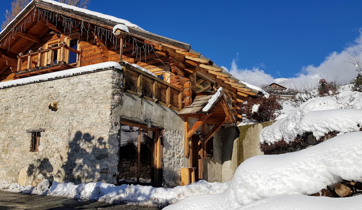 The exterior of an alpine chalet is displayed, featuring a combination of natural stone and wooden accents. Snow covers the roof and surrounding ground, creating a serene winter scene. Clear blue skies complement the rustic charm of this mountain retreat.