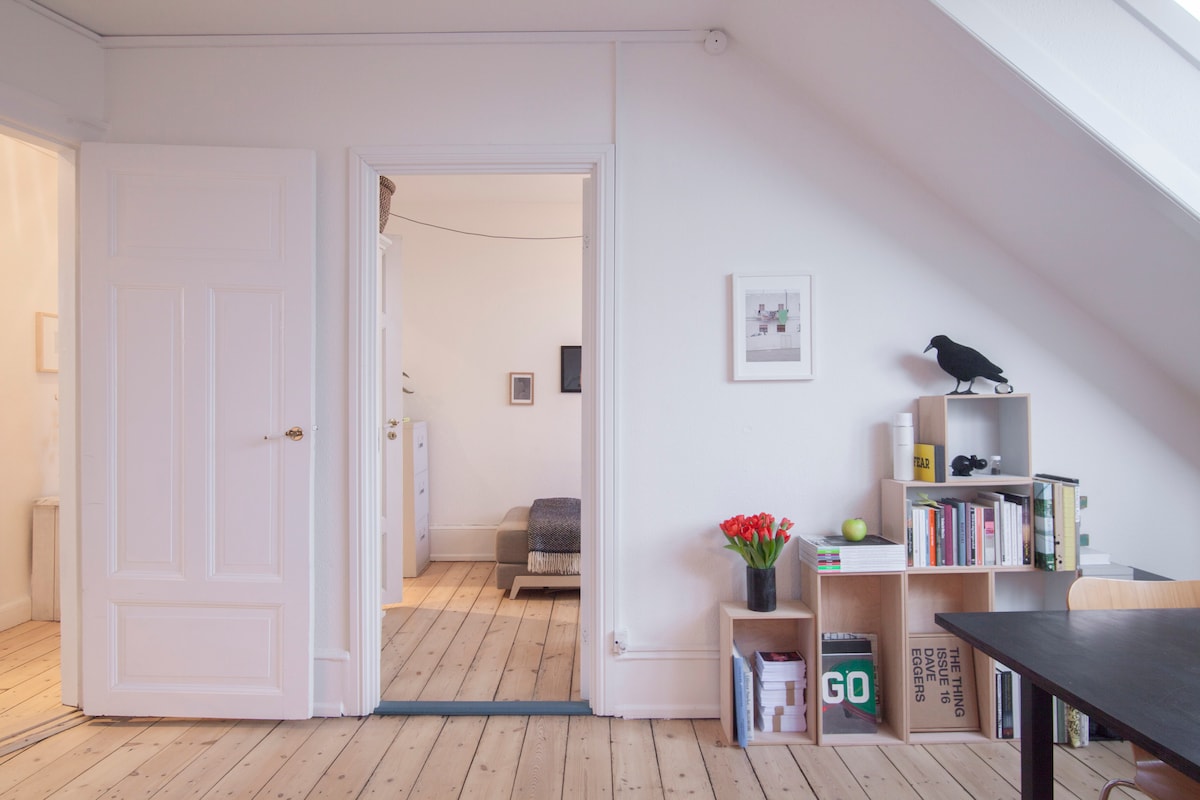 A bright interior space features white walls and a wooden floor, creating an open and airy feel. A small bookshelf, adorned with books and a vase of flowers, is positioned beside a dining table. Doorways lead to additional rooms, hinting at the loft's spacious layout.