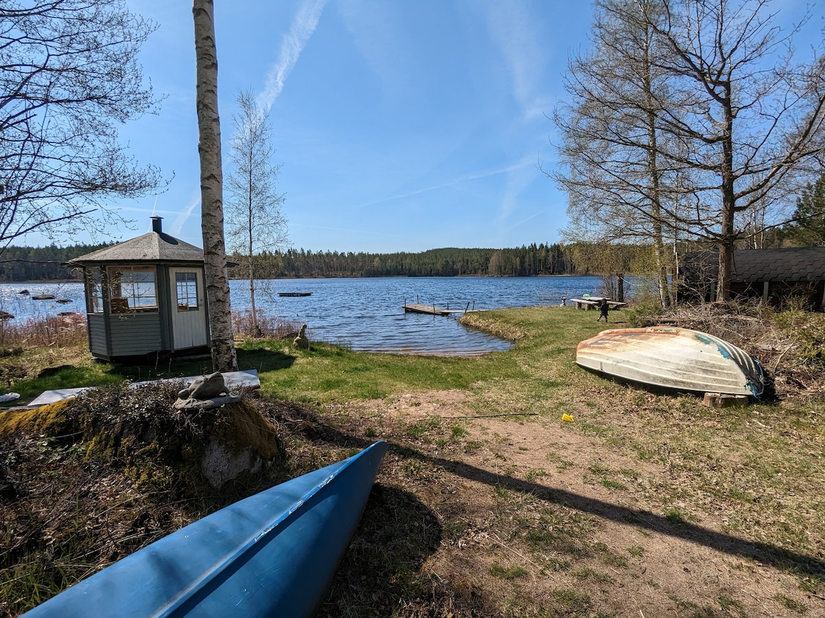 A serene view of a lake is visible, with a small gazebo and several boats along the shore. Lush green grass meets the sandy beach, leading to the water's edge. Surrounding trees provide a natural backdrop against a clear blue sky.