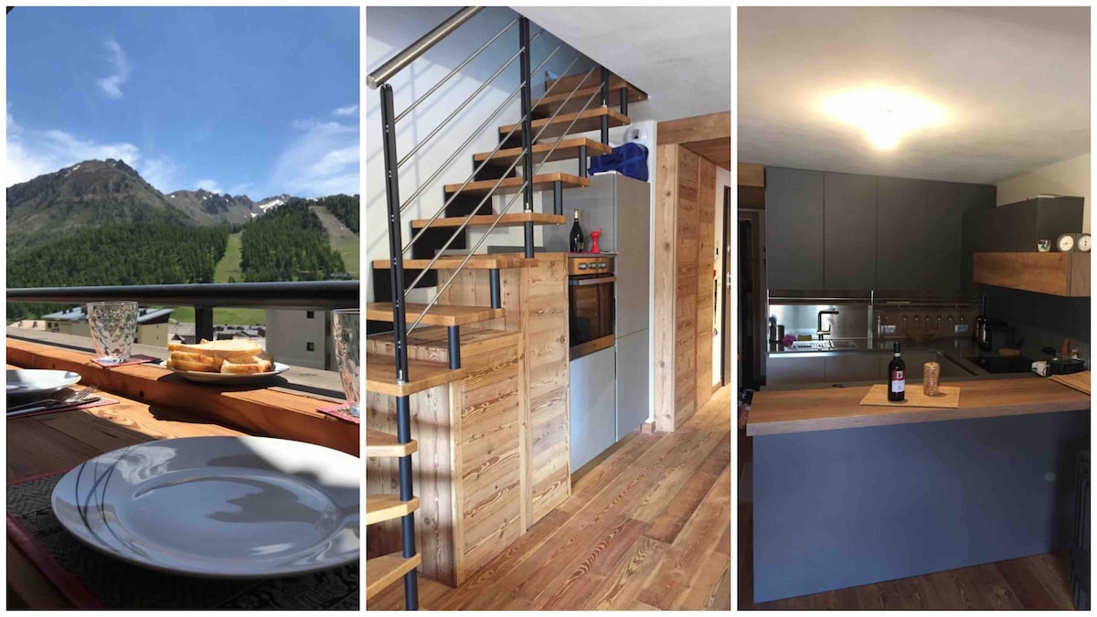 A balcony table is set for dining, featuring plates and glassware against a backdrop of majestic mountain views. The spacious wooden staircase leads to upper living areas. A modern kitchen showcases sleek cabinetry and appliances, with wooden accents adding warmth to the design.