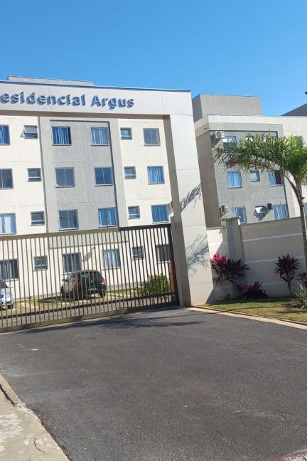 The image displays the front exterior of Residencial Argus, characterized by a modern building structure with multiple windows. A gated entrance is visible, featuring a paved driveway that leads to the facility. Lush green plants and decorative shrubs are positioned along the entrance area.