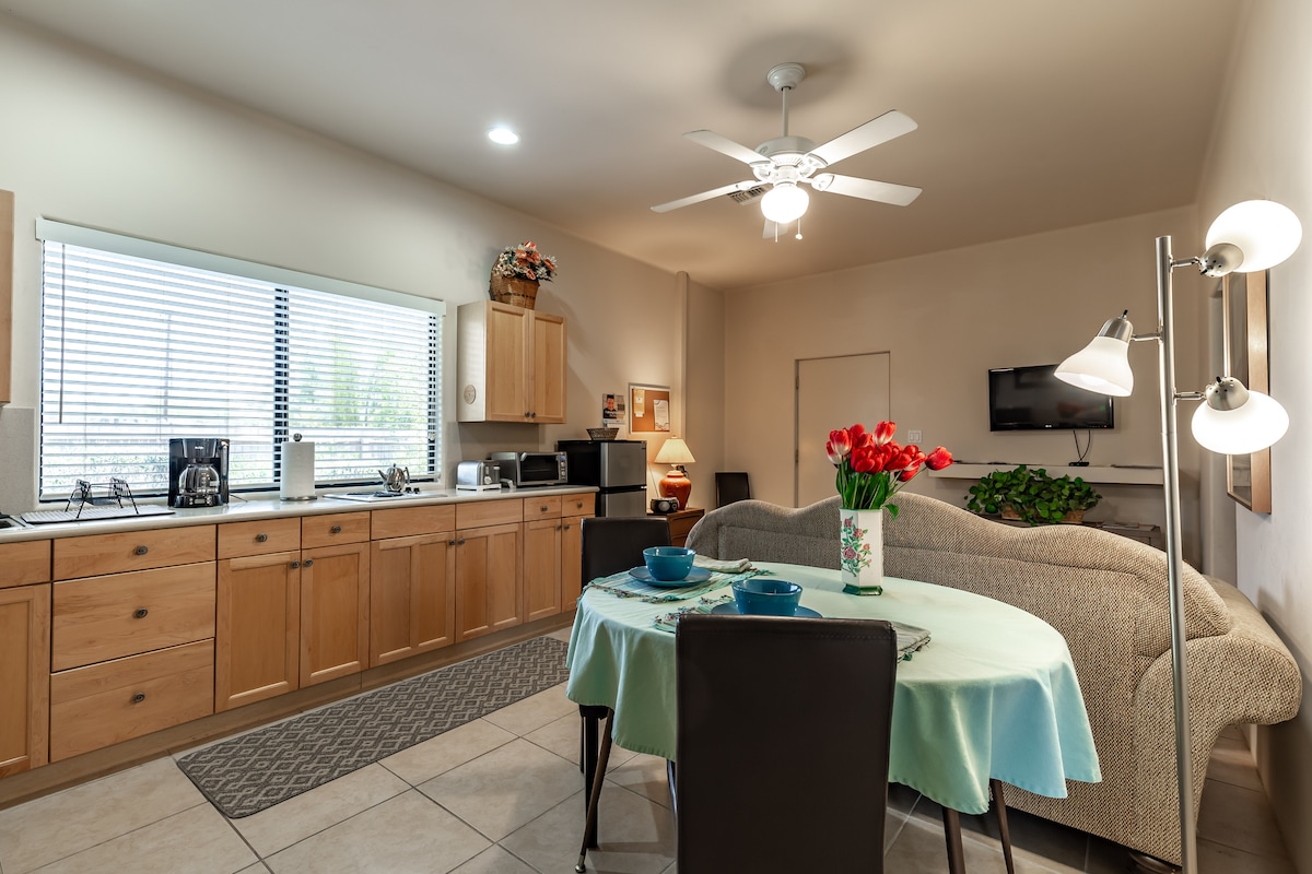 A bright kitchen area is presented with wooden cabinetry and a compact dining space. A round table is set with blue dinnerware, complemented by a vase of red tulips. A ceiling fan circulates air above, with natural light filtering through the window.