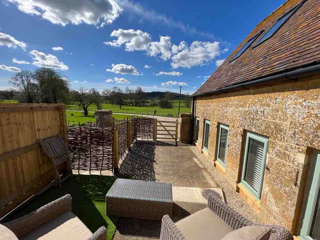 A patio area is visible, featuring two comfortable seating chairs and a small table. The space offers scenic views of the surrounding green fields and trees under a clear blue sky with fluffy white clouds.