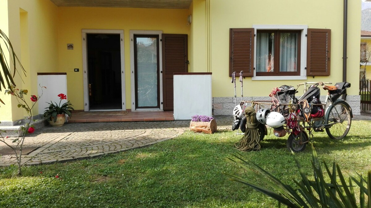 The exterior of the ground-floor apartment is showcased, featuring a sunlit garden area with a well-kept lawn. A parked bicycle with gear and accessories is positioned nearby, along with climbing equipment. The entrance is flanked by large windows and wooden shutters.