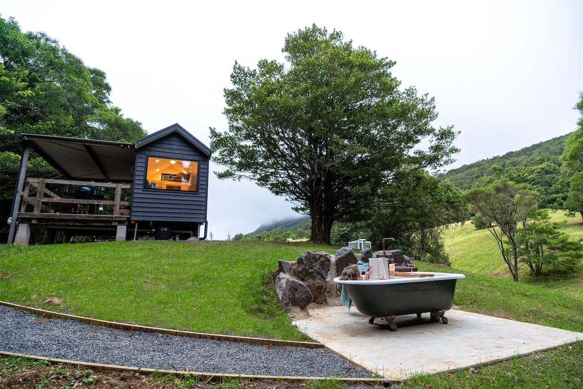 The tiny house is set within a lush green landscape, bordered by trees and rolling hills. An outdoor bath is situated on a stone pad, complemented by natural rock formations. The covered deck features large windows, offering views of the serene surrounding nature.