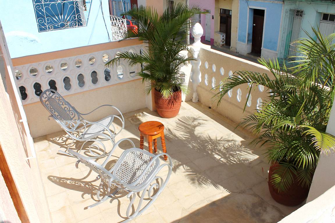 A spacious terrace is featured, adorned with two metal lounge chairs and a small wooden table. Potted plants add a touch of greenery, while the sunlight casts soft shadows on the tiled flooring. The surrounding railings provide a sense of openness to the outside.