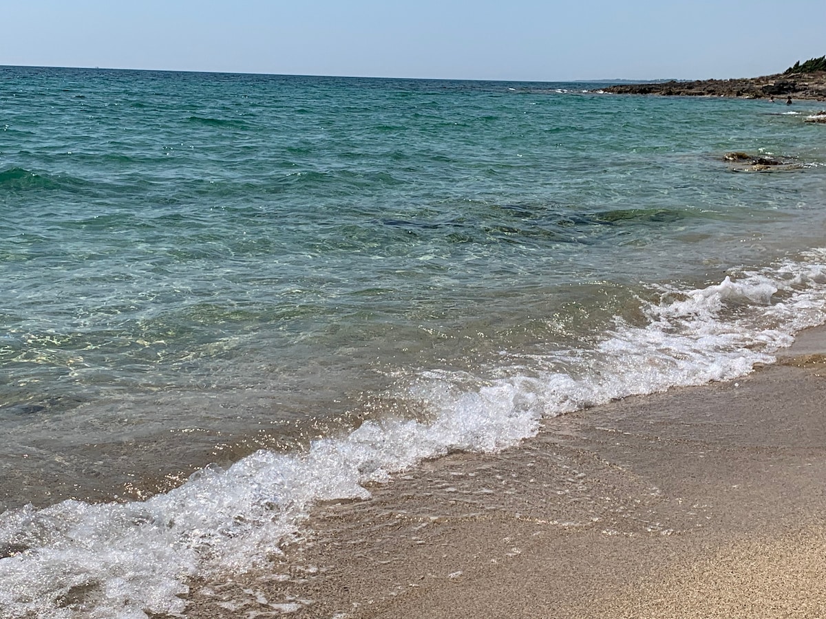 A clear view of a beach is shown, with gentle waves lapping at the sandy shore. The water appears calm, featuring shades of blue and green. In the distance, rocky formations are visible, enhancing the coastal landscape.