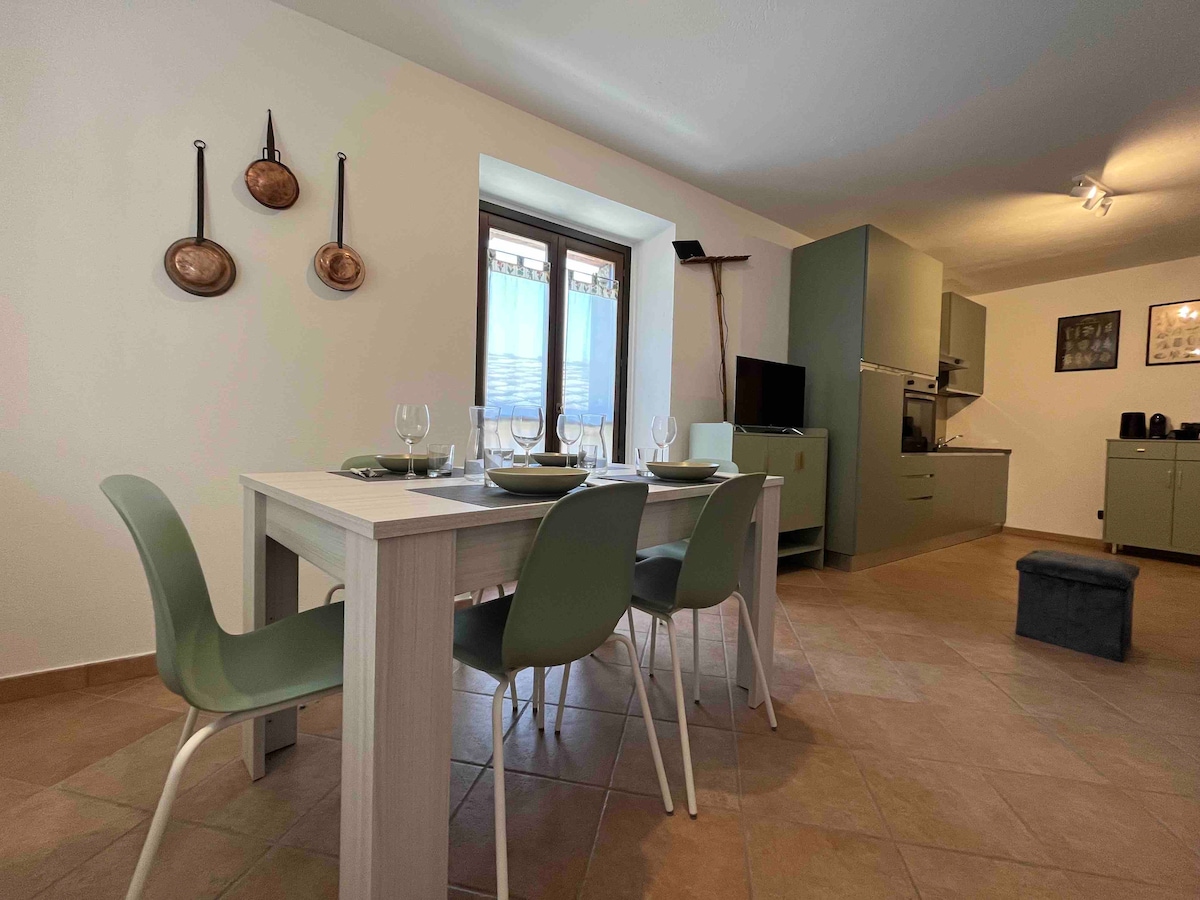 A dining area features a light wooden table with seated chairs, set up for a meal with plates and glasses. A window allows natural light to fill the space, with a view of the outside. The kitchen area is visible in the background, showcasing ample cabinetry.