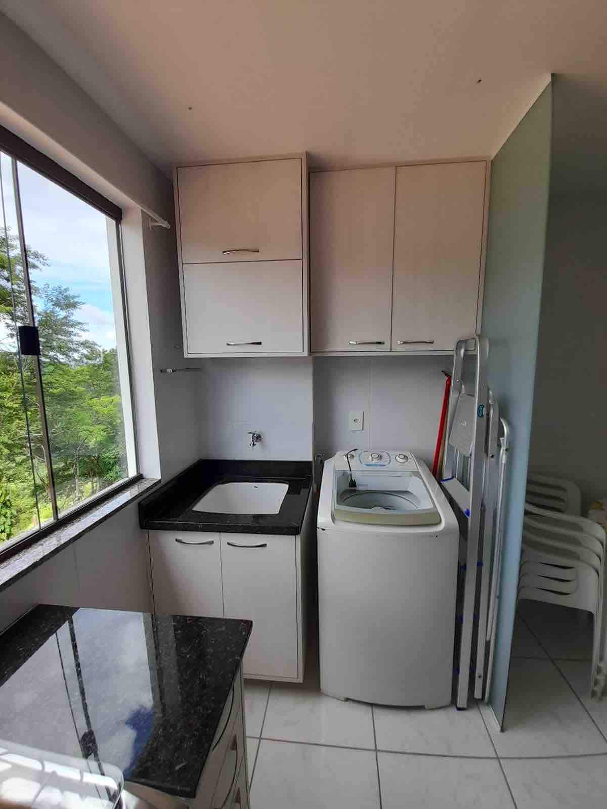 A compact kitchen area is visible, featuring a modern countertop and cabinets. A washing machine is positioned next to the sink, with a window providing a view of greenery outside. Stacked plastic chairs are also noted in the corner, adding to the functional space.