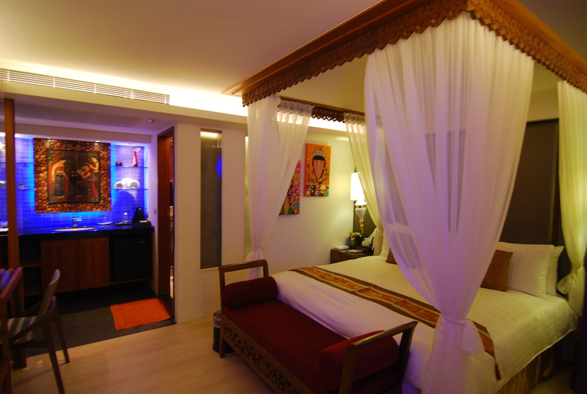 A comfortable sleeping area is framed by elegant drapery, with a spacious bed featuring decorative headboards. Soft lighting creates a warm ambiance, while vibrant artwork adorns the walls. A kitchenette is visible in the background, complemented by a small dining area.