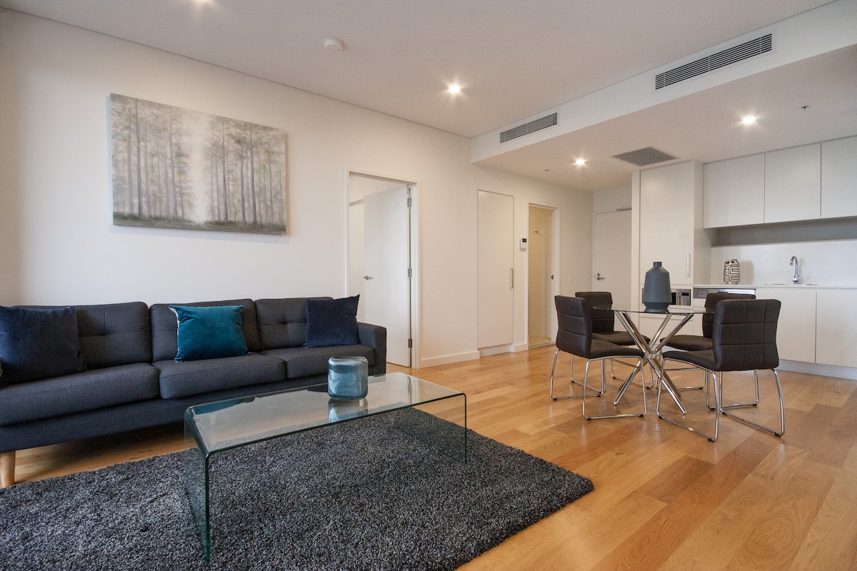 A modern living space features a dark sofa paired with accent pillows. A glass coffee table rests upon a soft rug, while a dining area with four chairs is visible. Natural light fills the room, highlighting the sleek kitchen in the background.