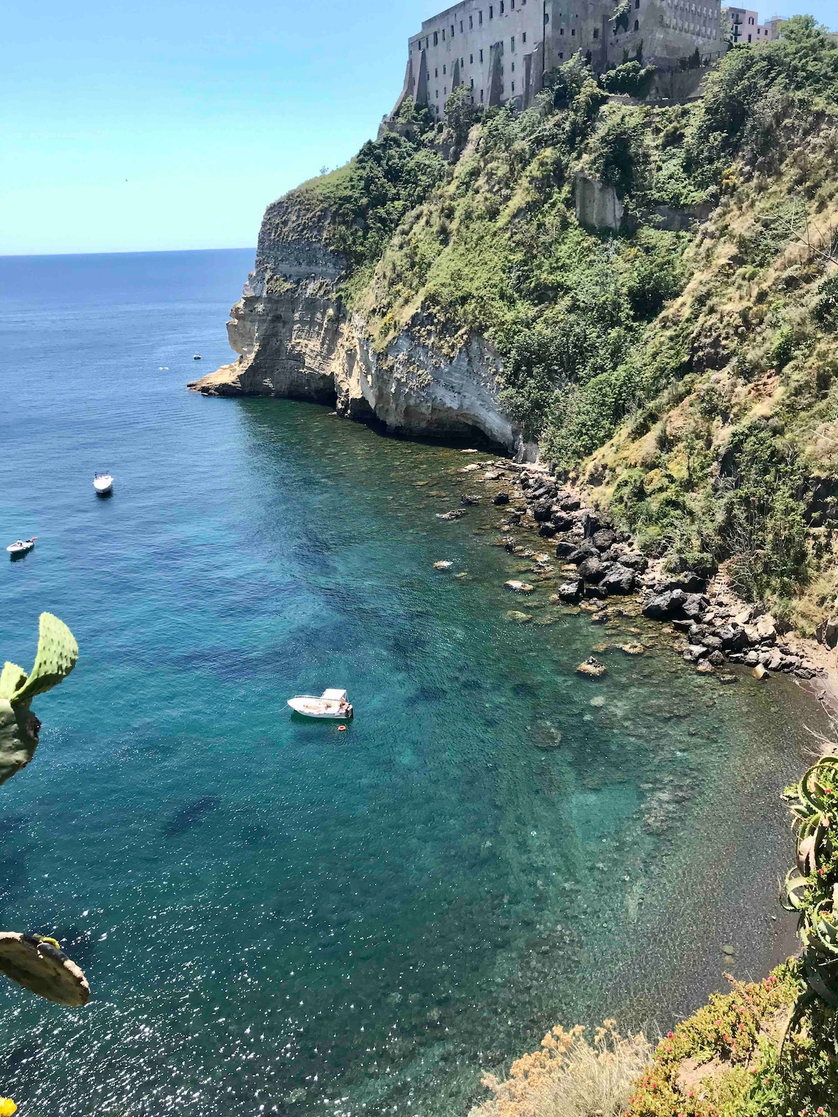 A serene coastal scene showcases clear blue waters gently lapping against rocky shores. A small boat can be seen navigating the tranquil waters, framed by lush greenery and the rugged coastline. A historic building rises prominently above, adding character to the natural landscape.