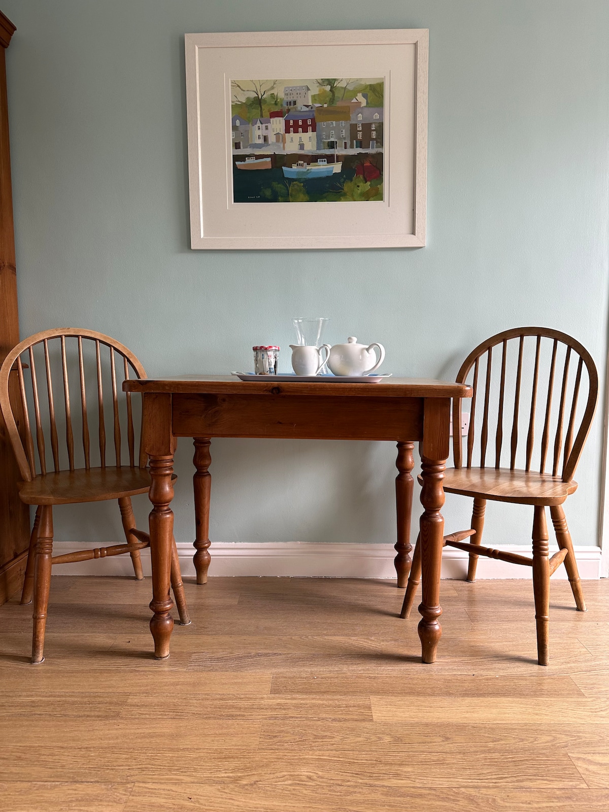 A small, wooden table with turned legs is positioned between two classic spindle-back chairs. A framed artwork hangs on the wall above, featuring a serene landscape with buildings and greenery. Soft light illuminates the light wood floor, creating a welcoming atmosphere.