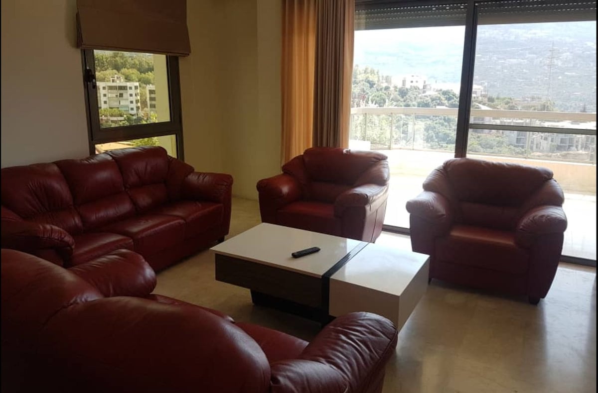 A comfortable seating area is arranged with two large sofas and two armchairs, all in rich red leather. A modern coffee table sits at the center, and large windows provide natural light and a view of the surrounding hills.