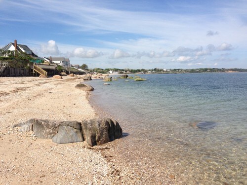A tranquil beach scene displays a gentle coastline with smooth stones partially submerged in clear water. Soft sand and sparse pebbles line the shore, while low, green vegetation is visible in the background. A bright sky with scattered clouds enhances the serene atmosphere.