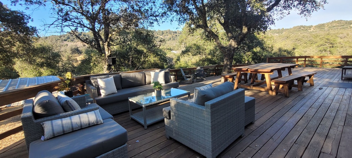 An expansive wooden deck is furnished with a comfortable seating area featuring a low table surrounded by woven chairs, inviting relaxation. A large picnic table offers ample space for gatherings. The surrounding oak trees enhance the serene outdoor environment.
