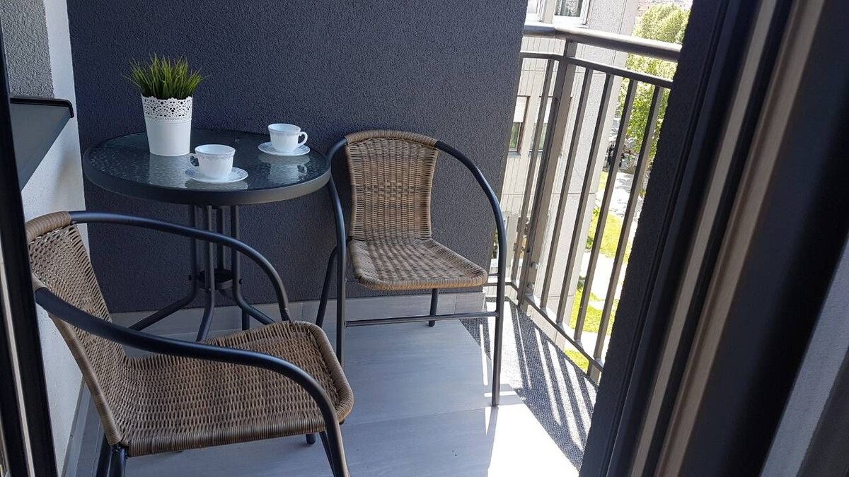 A small terrace features a round table accompanied by two chairs. A potted plant decorates the table, while a tea set is placed on it. The surrounding area includes a view of the exterior of the building and greenery.