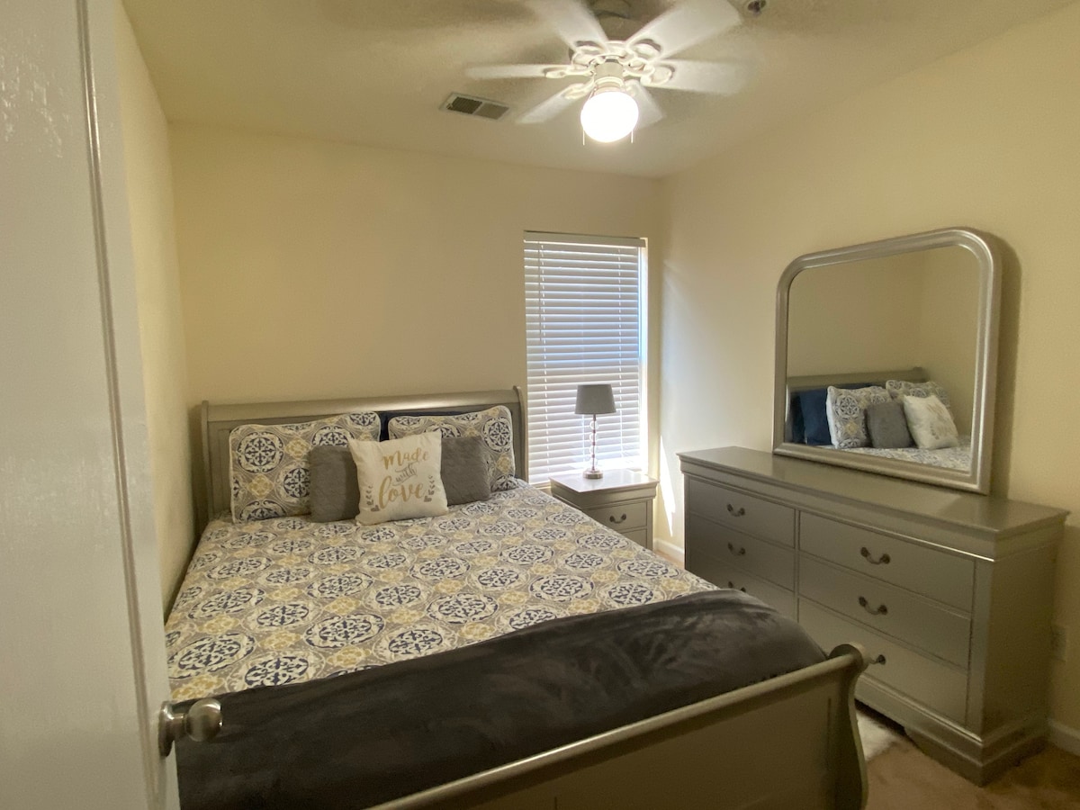 A cozy bedroom features a spacious bed with patterned bedding and decorative pillows. A dresser and a mirror are positioned along one wall, while a bedside lamp sits on a small nightstand next to the window, which allows natural light to filter into the space.