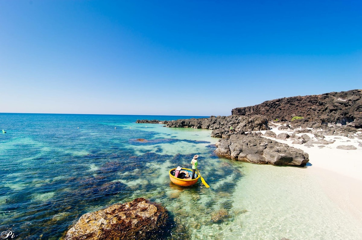 A serene beach scene features clear turquoise waters gently lapping against smooth rocks. A small canoe is observed near the shore, where sand meets water. The horizon displays a vibrant blue sky, enhancing the peaceful atmosphere of this coastal landscape.