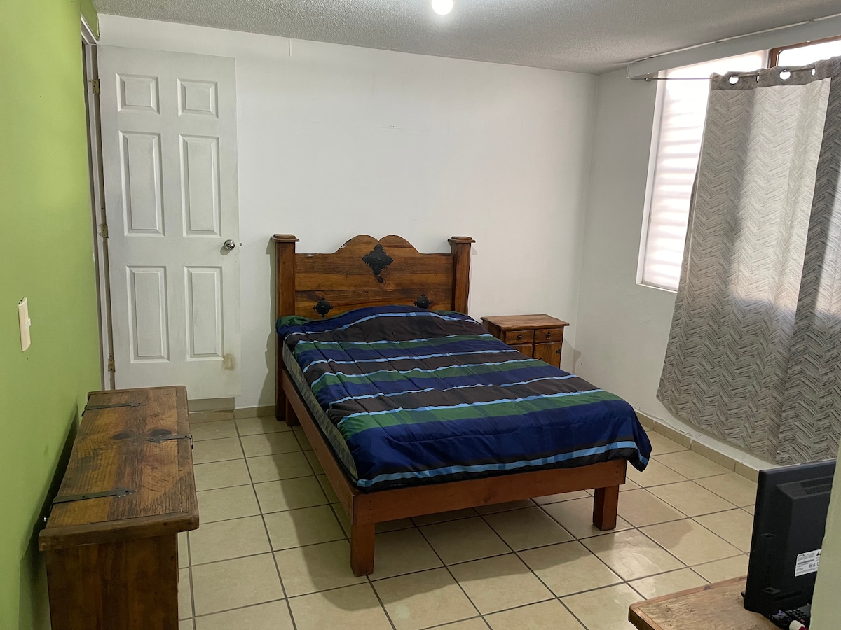 A bedroom is presented with a spacious wooden bed featuring a striped blue and green blanket. Two wooden nightstands sit beside the bed. Light enters through a nearby window, which is framed by light grey curtains, while a small television is placed on a wooden dresser.