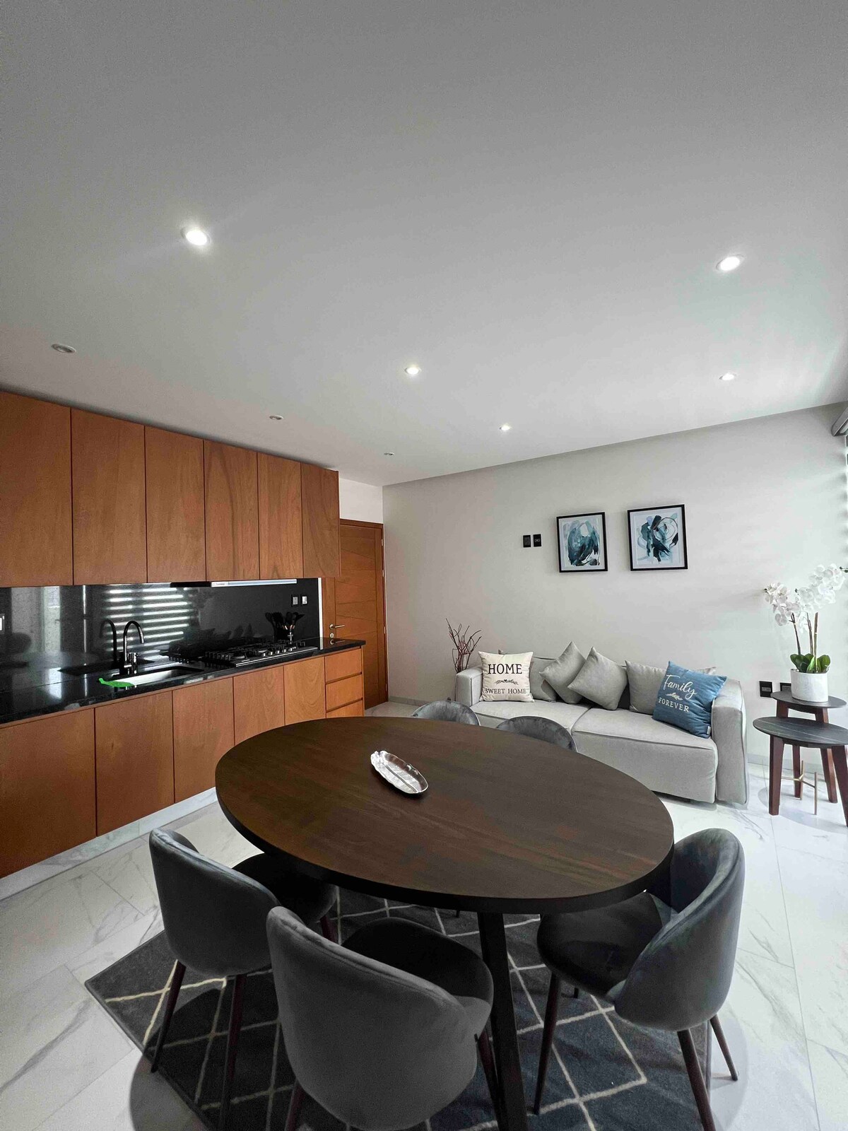A modern living area is showcased, featuring a round dining table surrounded by plush chairs. A comfortable sofa is positioned against the wall, complemented by framed artwork. The space is well-lit with recessed ceiling lights, and a sleek kitchen area is visible in the background.