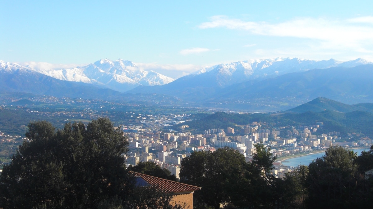 A panoramic view of the city of Ajaccio is presented against a backdrop of snow-capped mountains. Lush greenery and hills frame the scene, while the coastline and clear skies enhance the natural landscape. The urban area shows a spread of buildings nestled among the hills.