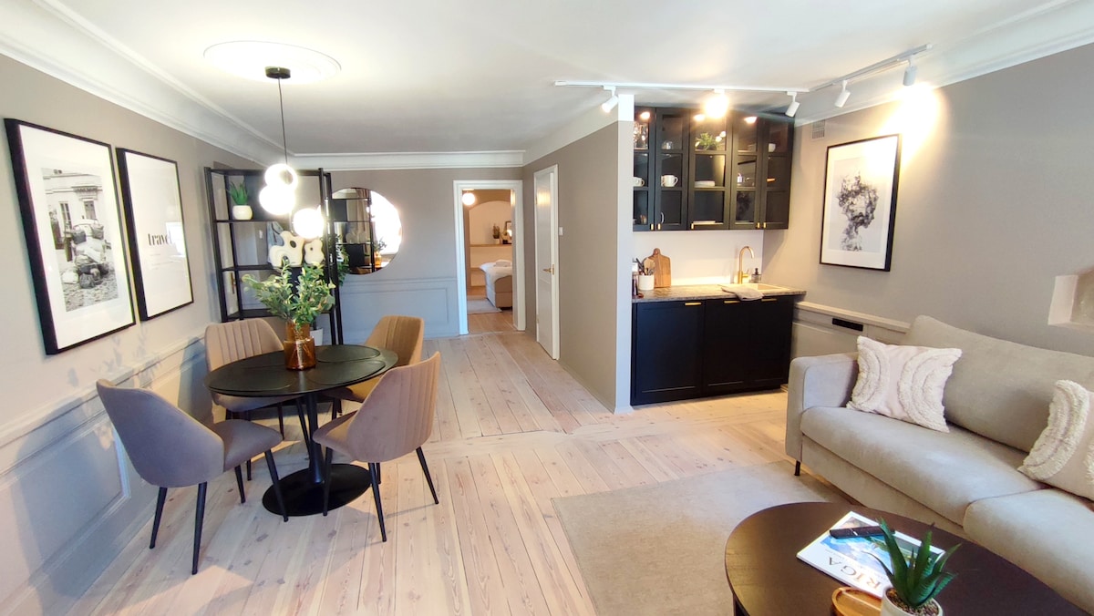 A spacious living area is highlighted by a dining table with four chairs and a comfortable sofa. Dark cabinets line the back wall above a modern kitchen countertop. Natural light illuminates the wood flooring, creating a welcoming atmosphere with framed artworks on the walls.