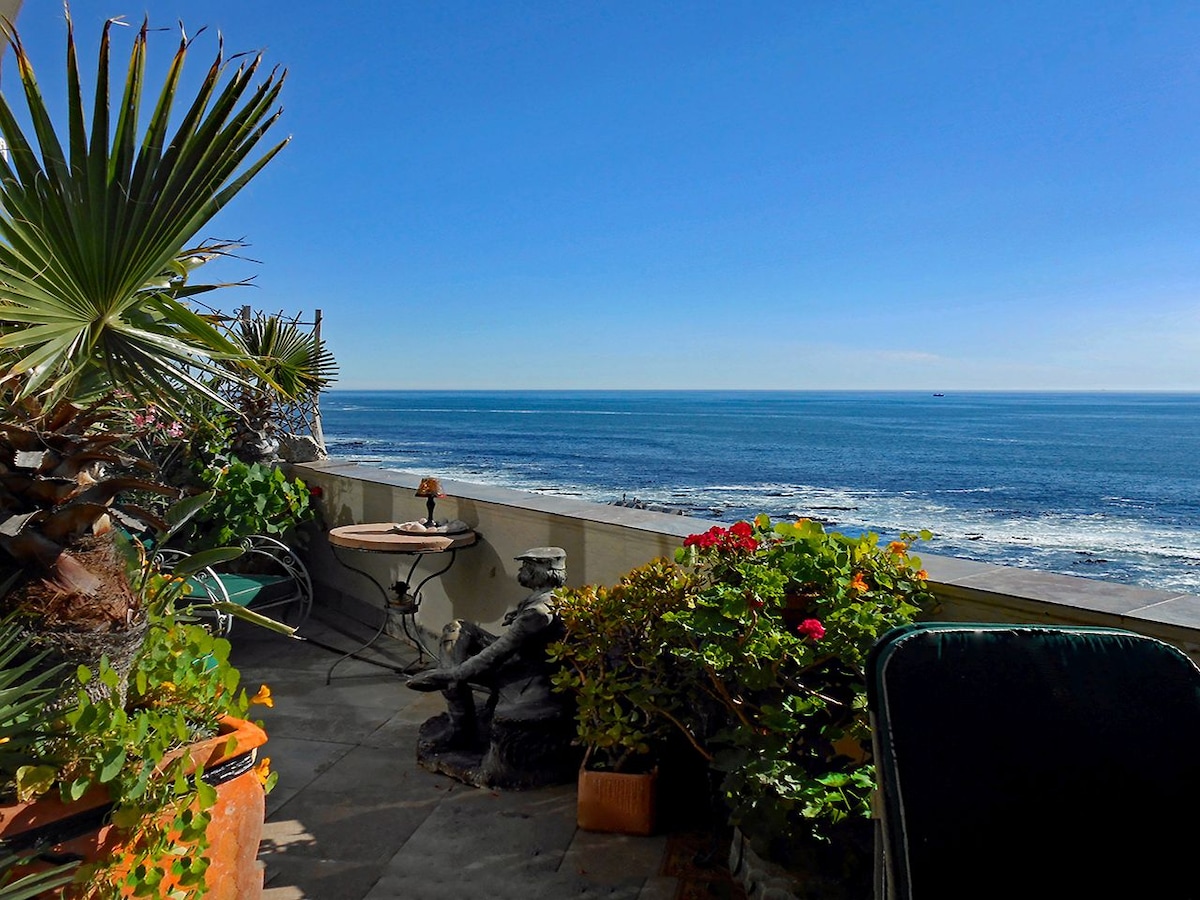 An outdoor balcony offers an unobstructed view of the ocean, framed by lush greenery and colorful flowers in terracotta pots. A small table rests beside a decorative statue, providing a tranquil space to enjoy the surrounding scenery.