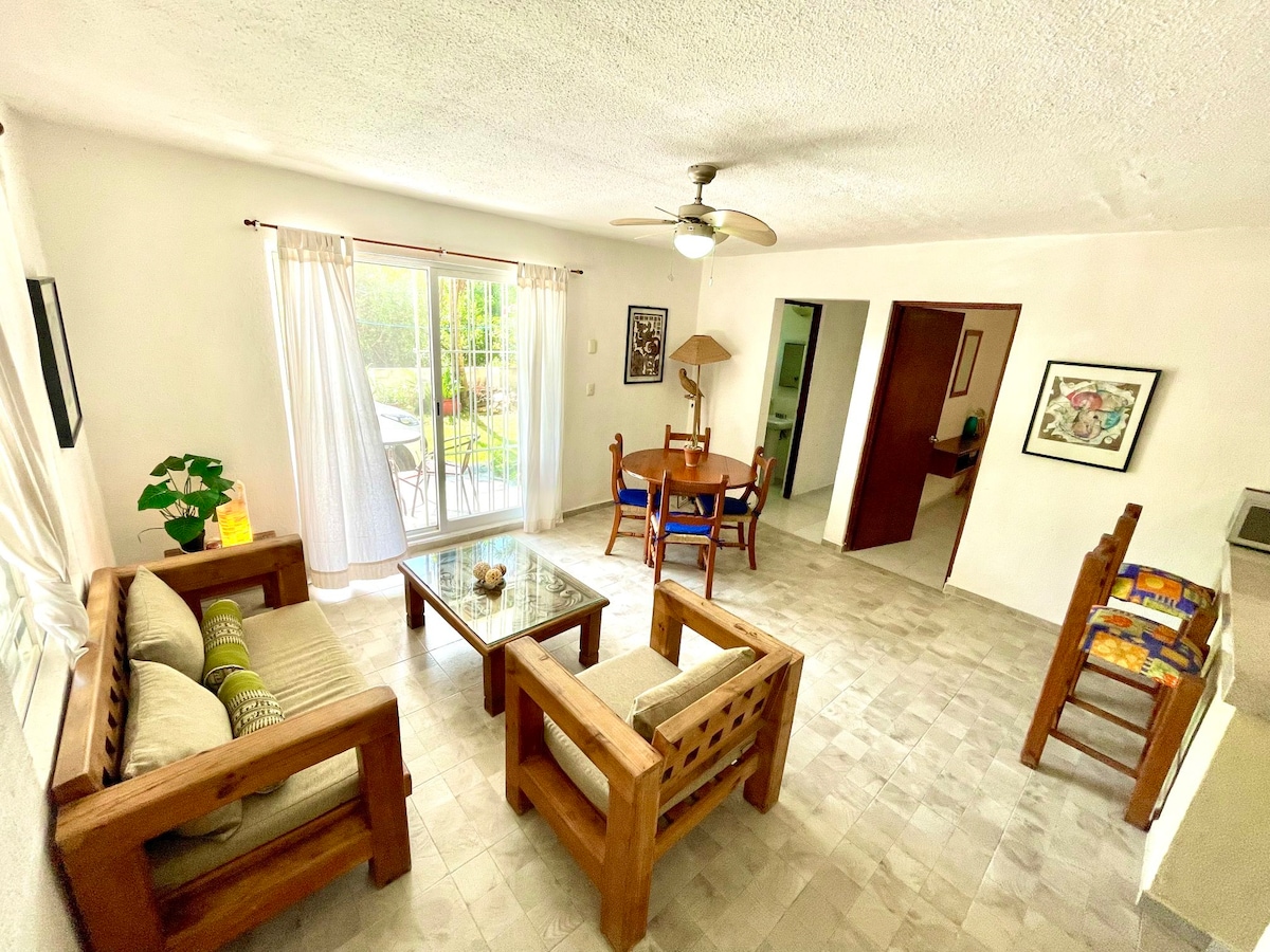 A spacious living area features two wooden sofas adorned with decorative cushions, positioned around a glass coffee table. Natural light floods in through large sliding doors, which provide access to a garden terrace. A dining table for four is visible in the background.