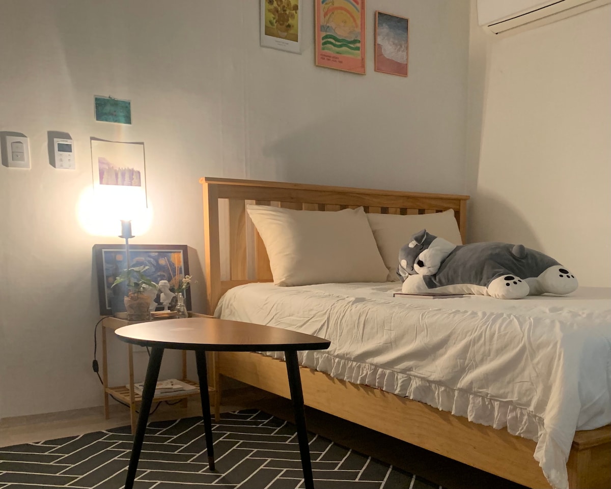 A cozy bedroom features a wooden bed with soft linens and a plush whale-shaped pillow. A small round table is positioned nearby, accompanied by a lamp that provides warm lighting. Wall art and decorative accents add a personal touch to the serene space.