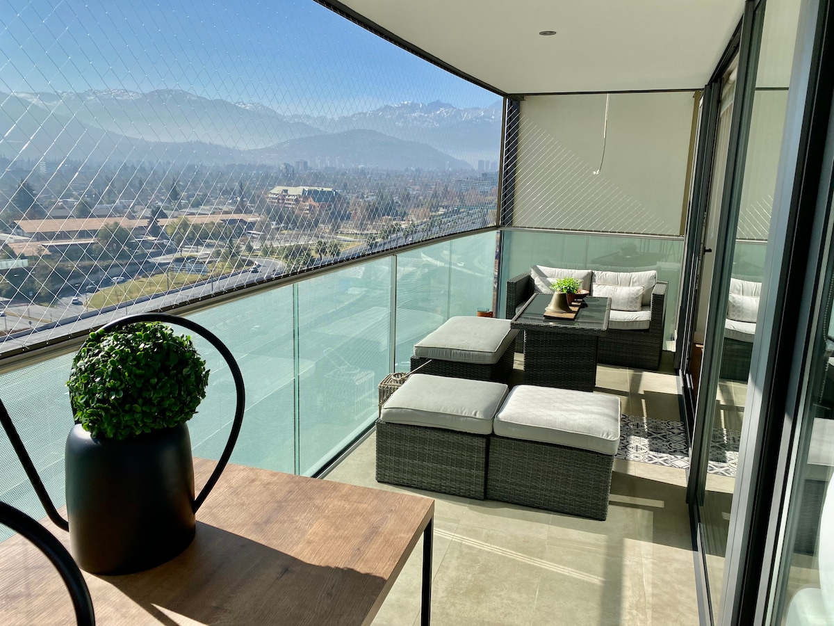 A spacious balcony features modern outdoor furniture arranged for relaxation, with a coffee table and plush seating. A decorative plant adds a touch of greenery. The expansive glass railing offers unobstructed views of the surrounding mountains and landscape, enhancing the tranquil atmosphere.