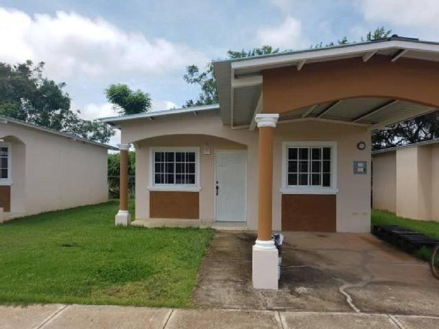 A single-story home is set within a green landscape, featuring a covered entrance with two columns. Large windows allow natural light to fill the space, while a well-maintained yard provides open greenery. Driveway space is visible, leading to the home's entrance.