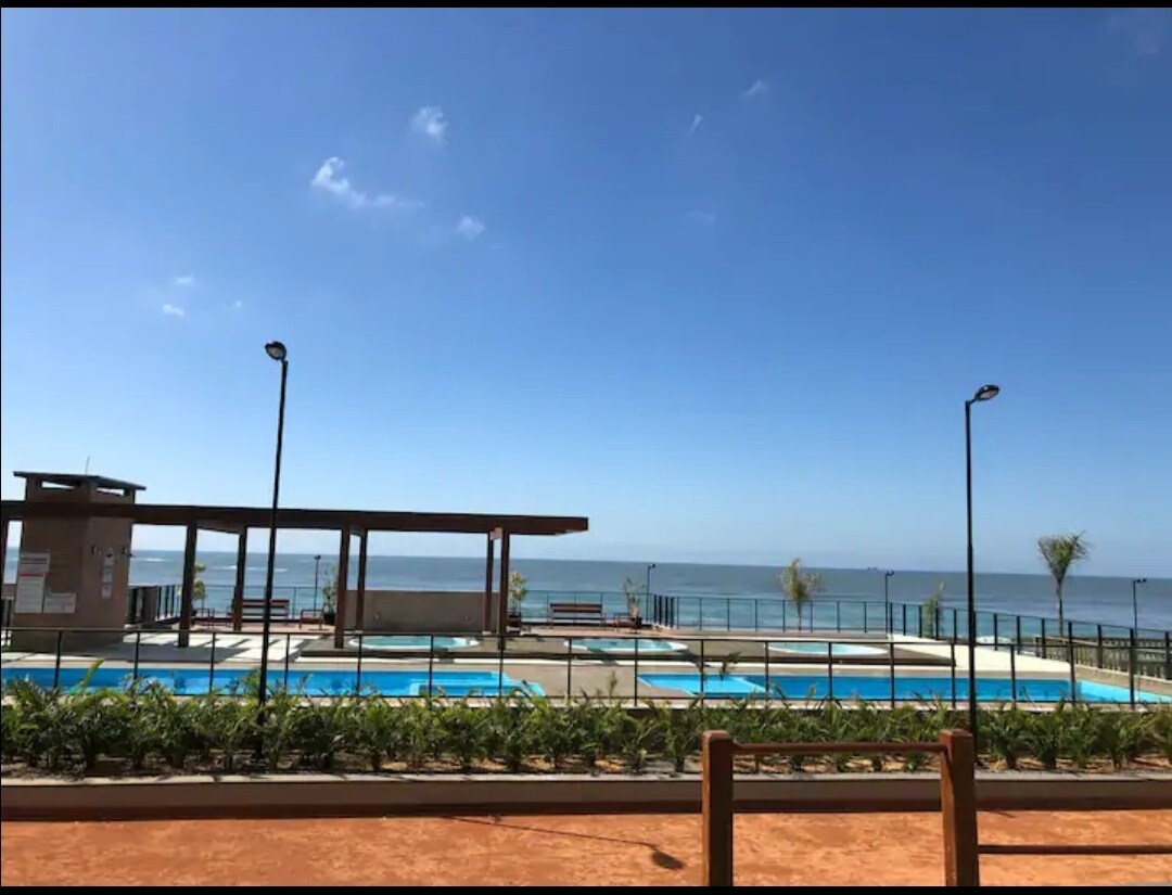 An outdoor area features several swimming pools bordered by palm trees. A modern pavilion with seating stands nearby, overlooking the expansive sea. The clear blue sky complements the serene atmosphere, enhancing the tranquil setting for relaxation.
