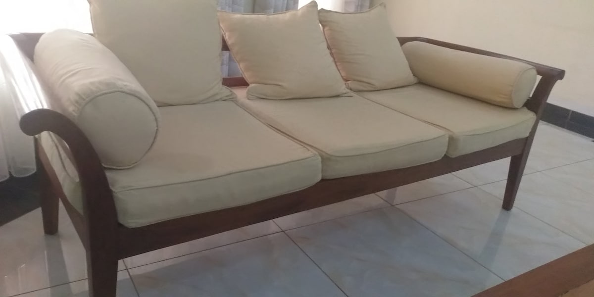 A wooden sofa with a simple design is displayed, featuring three spacious cushions in a light beige color. Two cylindrical pillows are placed at each end for added comfort. The sofa is positioned on a shiny tiled floor with natural light filtering through sheer curtains.