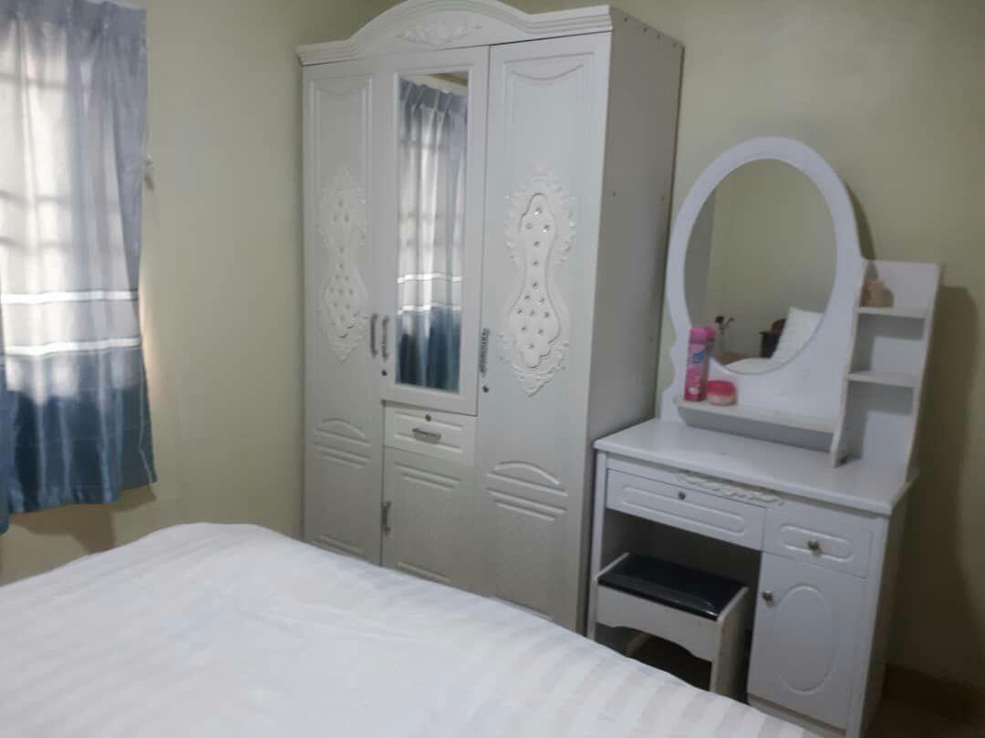 A light-colored bedroom features a spacious bed with subtle striped bedding. A white wardrobe with mirrored doors stands next to a vanity table with a round mirror. Soft blue curtains allow natural light to enter, enhancing the overall brightness of the space.