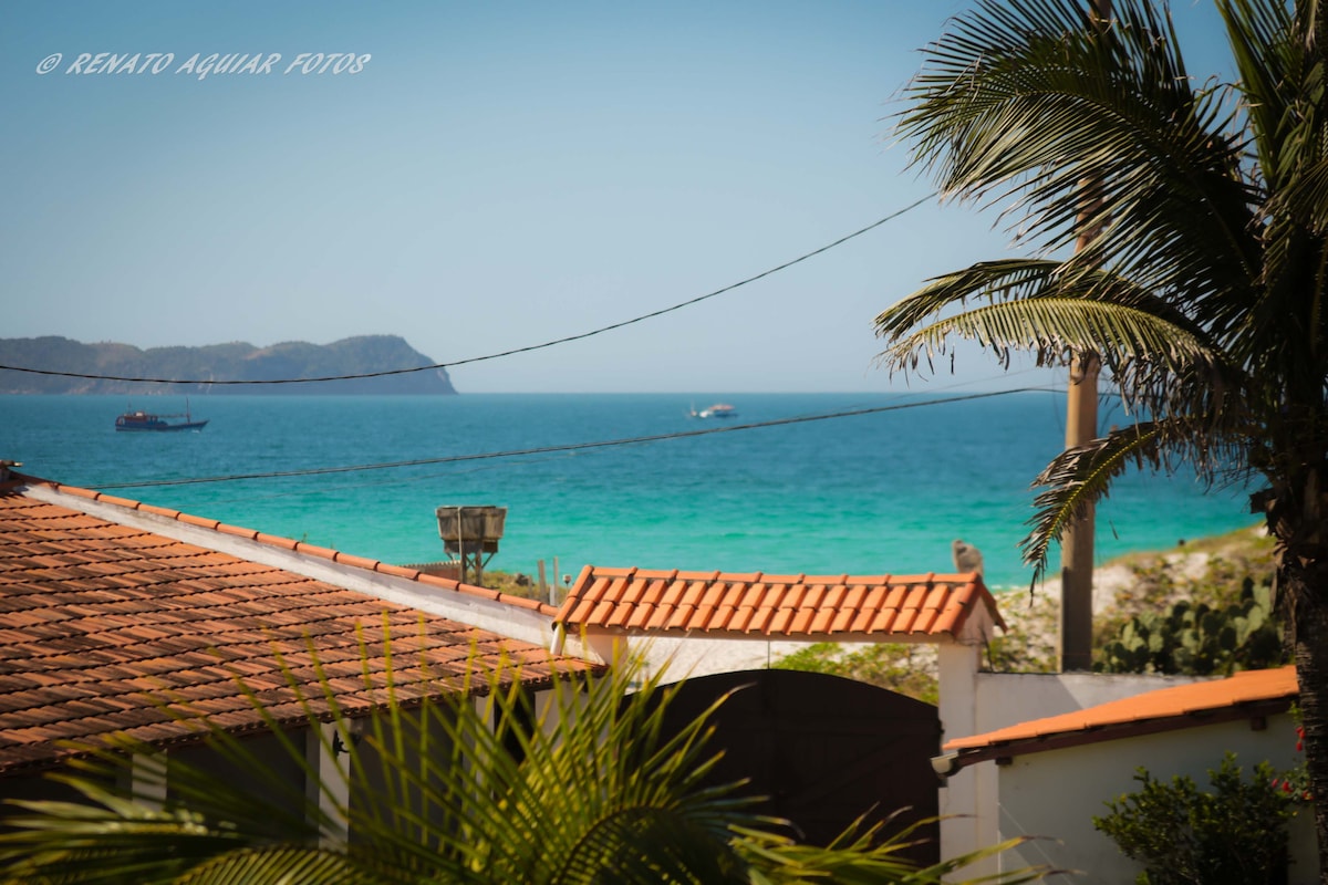 A view of the blue sea can be seen beyond a red-tiled roof, framed by palm leaves and green vegetation. Boats are visible on the water, with distant hills outlining the horizon.