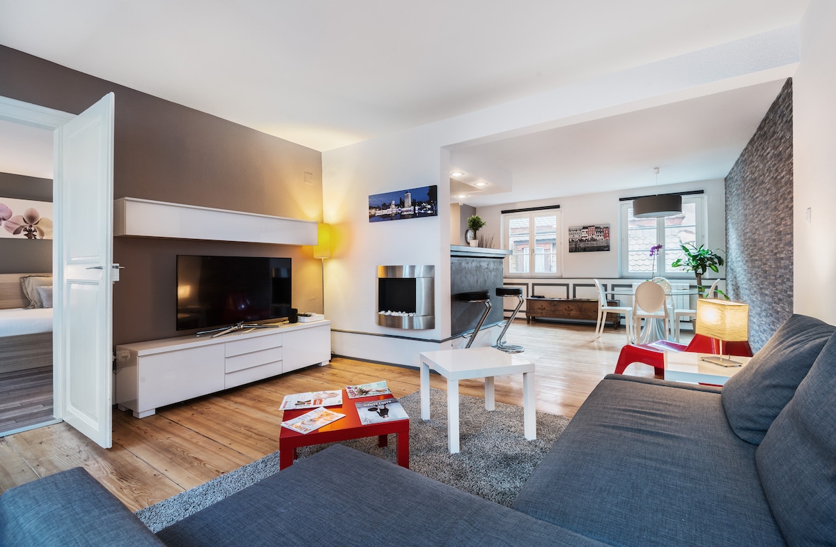 A modern living area is showcased with a gray sofa and a coffee table arranged on a textured rug. A flat-screen television is mounted on the wall, and a decorative fireplace adds warmth. Natural light fills the space through large windows, highlighting the wooden flooring.