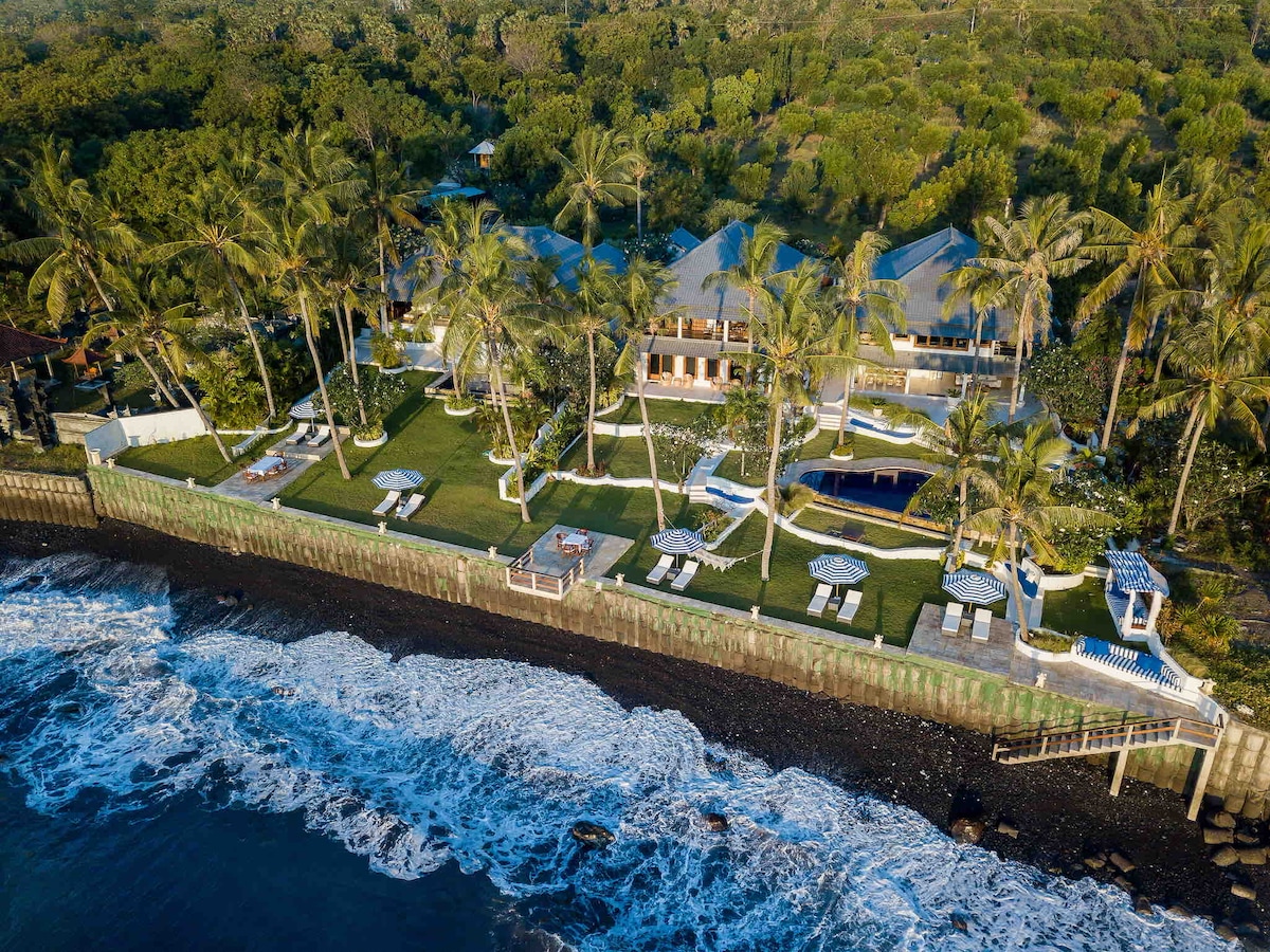 An aerial view captures the Kanna estate, showcasing spacious lawns bordered by palm trees, a pool area, and lounge chairs positioned beside the shoreline. The rhythmic waves of the Bali Sea create a serene coastal atmosphere, emphasizing the property's tranquil setting.