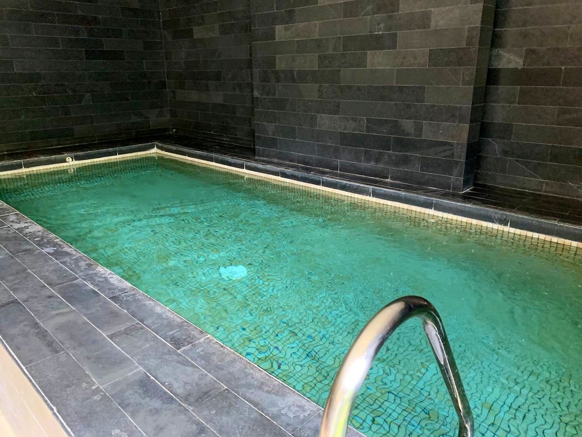 A sleek indoor pool is surrounded by dark stone walls, featuring a polished stone deck. A metal railing leads into the clear water, which reflects soft light, creating a serene swimming area. The overall design offers a modern and tranquil atmosphere.