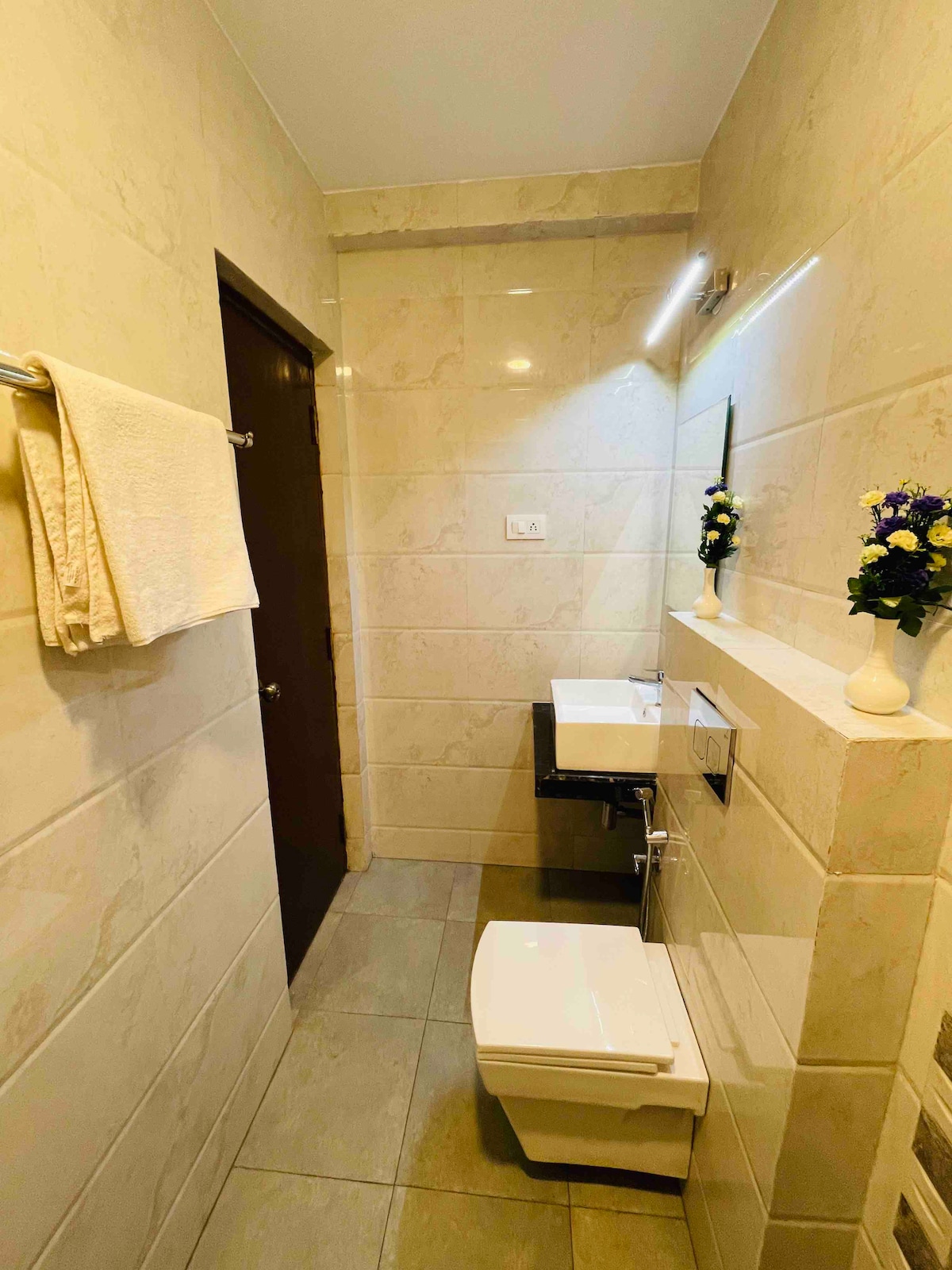 A compact bathroom is equipped with modern fixtures, including a sleek sink and a white toilet. Light-colored tiles cover the walls and floor, while a towel is neatly hung on a rack. Small decorative vases with flowers add a touch of color to the space.