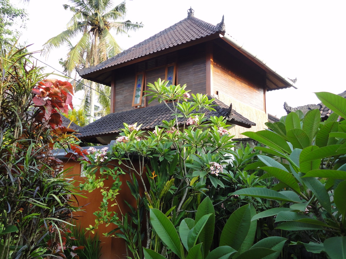 The two-story house is surrounded by lush tropical greenery and vibrant flowers. Its wooden facade features traditional Balinese architectural details, with a sloped roof adorned by intricate carvings. Sunlight filters through the foliage, highlighting the tranquil atmosphere of the property.