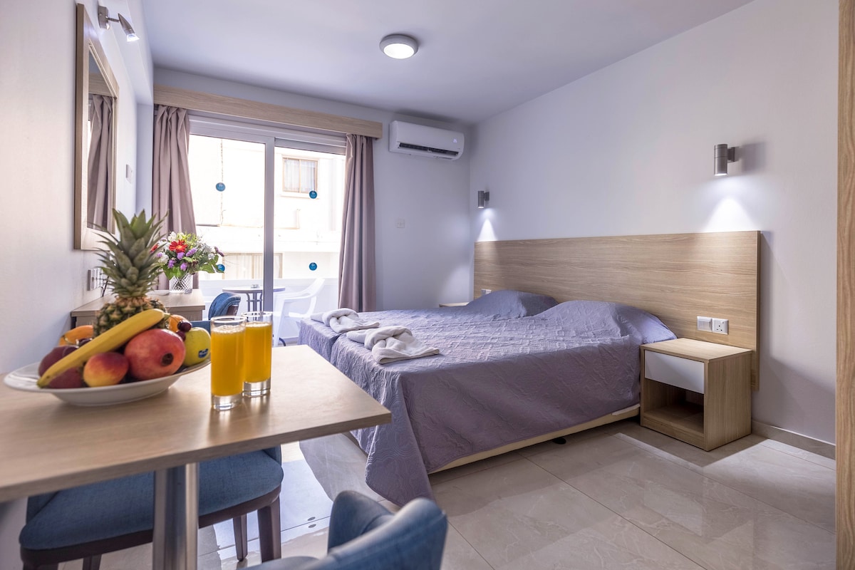 A well-lit room features a spacious bed with light bedding, accompanied by a bedside table. A small dining table holds a bowl of fresh fruit and two glasses of juice. Large glass doors lead to a private balcony, enhancing the airy ambiance.