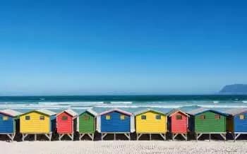 A row of colorful beach huts stands against a clear blue sky, aligned along the sandy shore. The vibrant hues of red, blue, yellow, and green create a striking contrast with the tranquil ocean waves gently lapping at the beach.