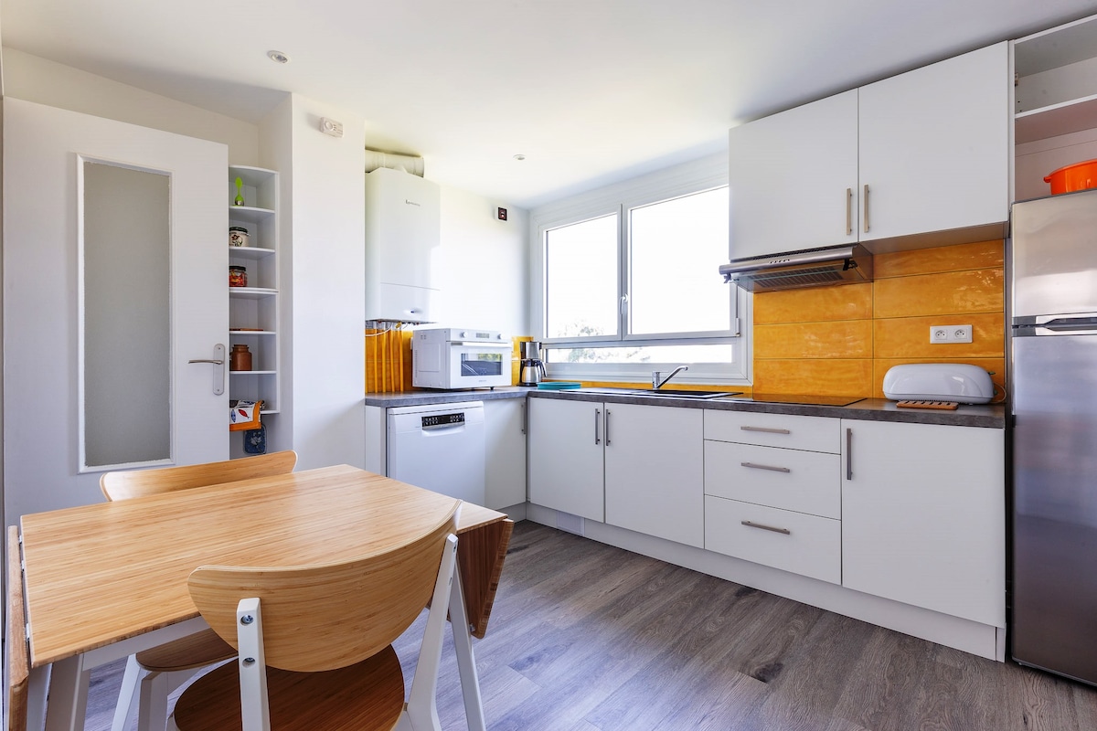 A modern kitchen is displayed, featuring white cabinetry and a bright orange backsplash. A wooden dining table with matching chairs is positioned nearby. Large windows allow natural light to illuminate the space, enhancing its functionality with appliances like a microwave and a refrigerator.