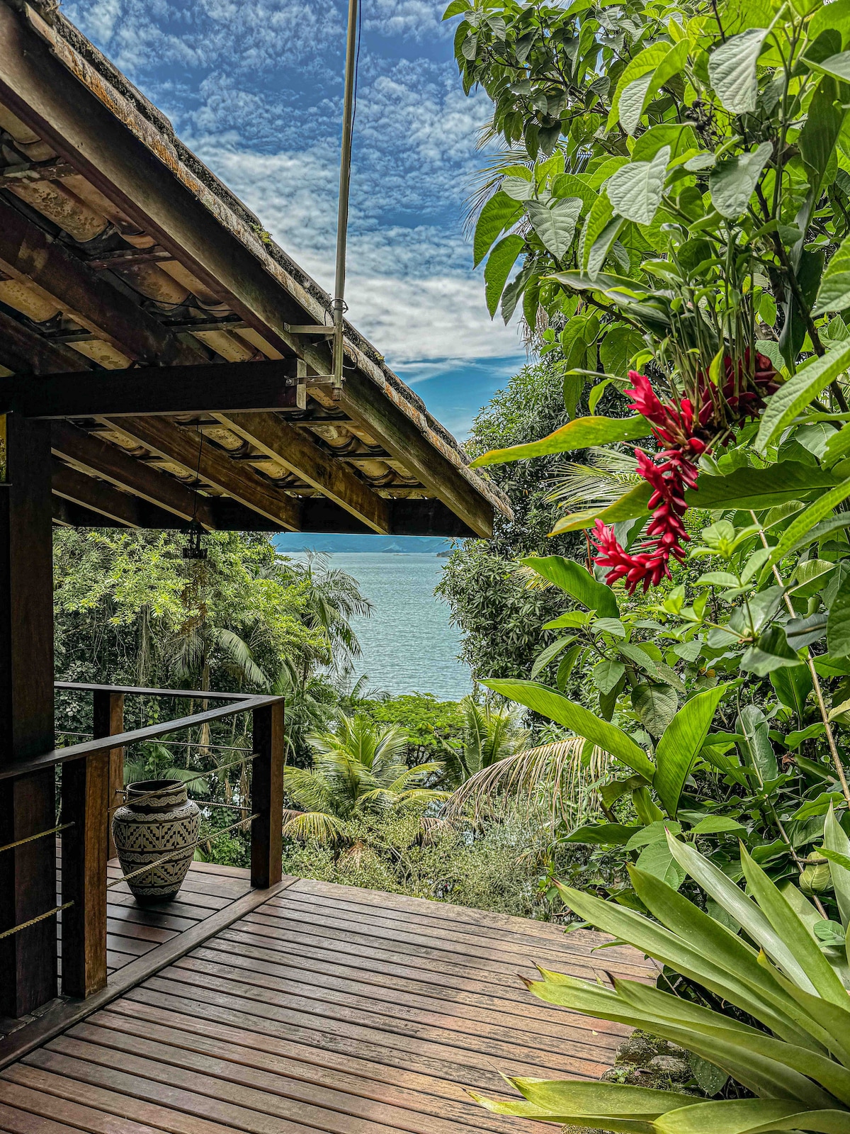 A wooden deck is seen overlooking lush greenery and the water. Tropical plants, including vibrant red flowers, frame the view, while the tranquil sea reflects the sky scattered with clouds. The rustic architecture of the house complements the natural surroundings.