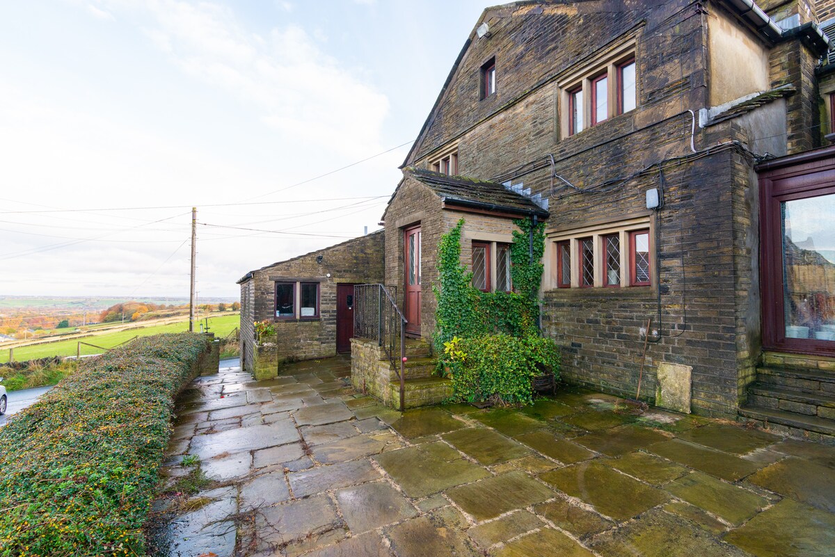 The exterior of a 400-year-old stone farmhouse is shown, featuring a rustic facade with a mix of stone and wood elements. A paved walkway leads to the entrance, bordered by greenery. Open fields are visible in the background under a partly cloudy sky.