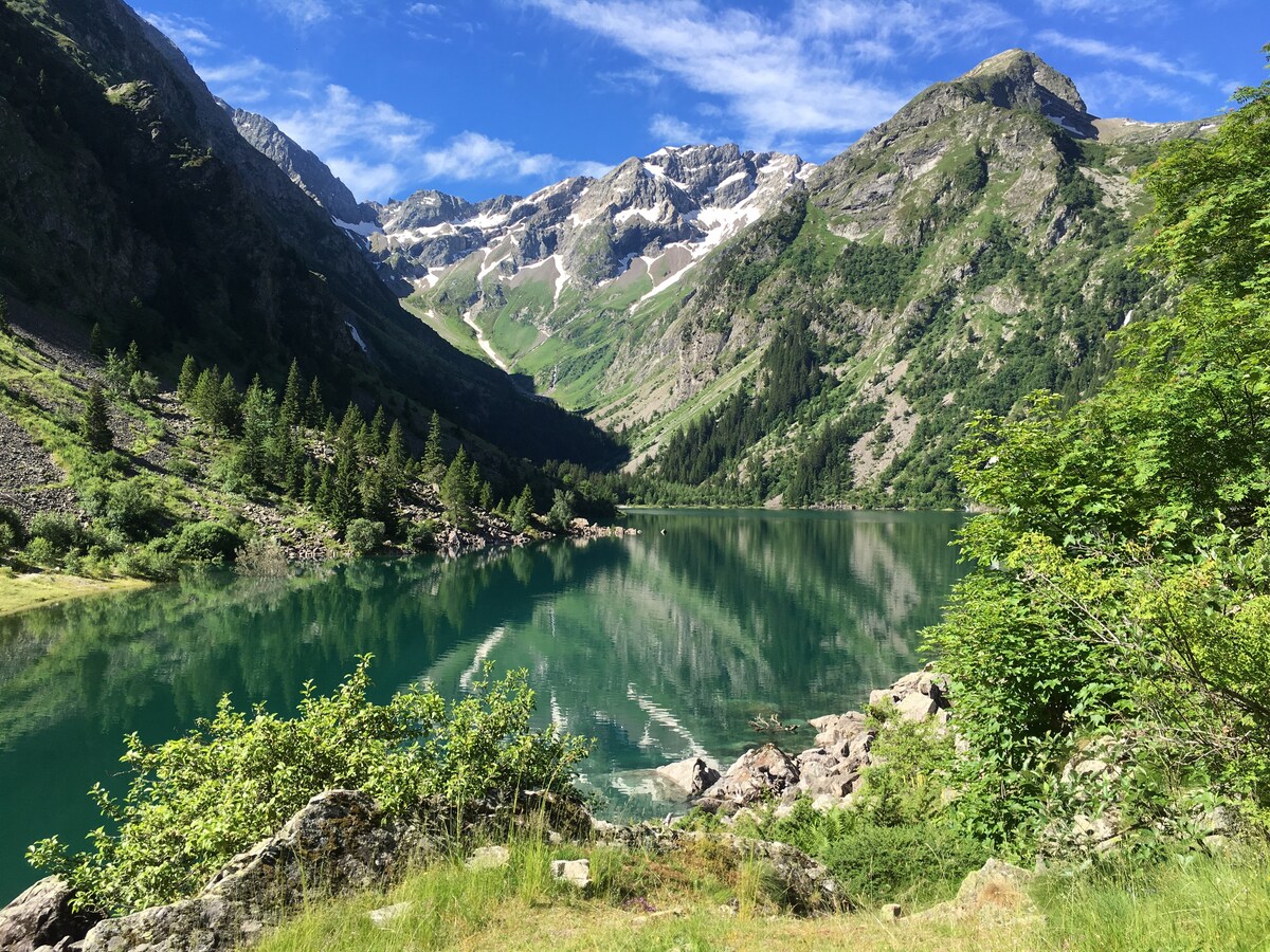 A tranquil lake is surrounded by majestic mountains, their peaks capped with patches of snow. Lush greenery lines the shore, reflecting beautifully in the calm water. Clear blue skies and gentle clouds complement the serene landscape, creating a peaceful and natural atmosphere.